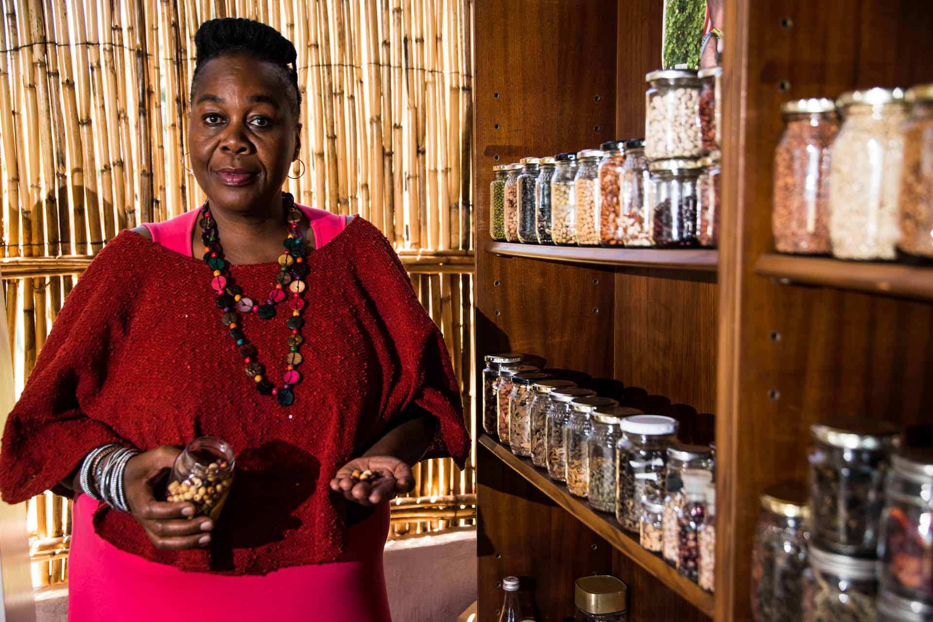 A foodie in a red sweater stands by shelves filled with jars of beans and grains. She holds an open jar in one hand, beans in the other, set against a warm bamboo background.