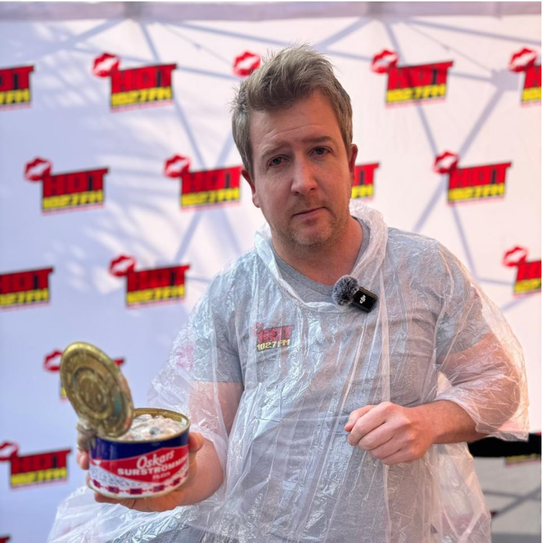 A man wearing a clear poncho holds an open can of surströmming, standing in front of a backdrop with repeating Hot logos. He appears to be preparing for a Teddython challenge or tasting.