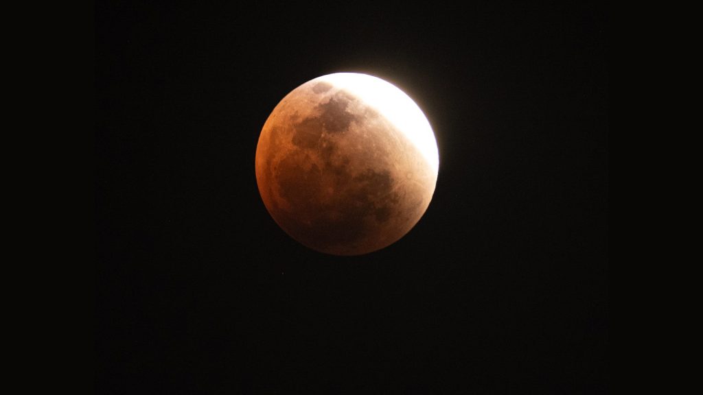 A partially eclipsed Blood Moon appears with a reddish hue and a bright white edge against the dark night sky, illustrating the progression of a lunar eclipse.