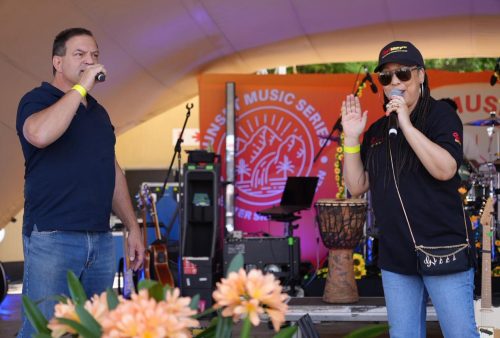 Two people, including a Dave Matthews tribute act, hold microphones and perform on an outdoor stage decorated with flowers and musical equipment. A bright orange banner reading Music Series is visible in the background.
