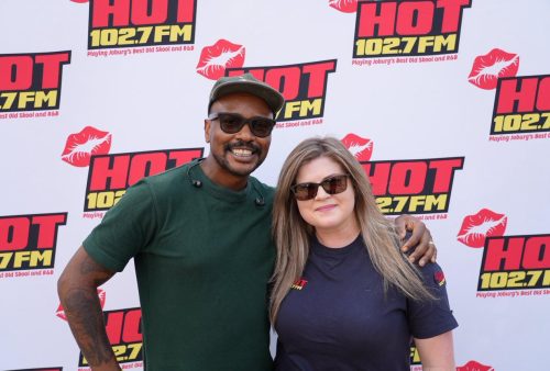 Two people stand smiling in front of a backdrop with the HOT 102.7 FM logo and red lips graphic, one in sunglasses and a green shirt, the other in a navy shirt—excited fans ready for the Dave Matthews event.