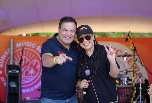 A man and a woman smile at the camera on stage. The man points while the woman, holding a microphone, flashes a peace sign. Both wear casual clothes and event hats. Sunset Music Series signage and Dave Matthews-themed gear appear in the background.