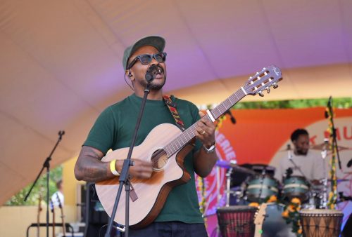 A musician wearing glasses and a green shirt plays an acoustic guitar and sings into a microphone on an outdoor stage, channeling Dave Matthews vibes. Another musician plays percussion in the background amid flower decorations.