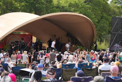 A large crowd watches musicians, including Dave Matthews, perform on an outdoor stage under a curved canopy, surrounded by trees. Many audience members are seated or standing, some wearing hats and colorful clothing.