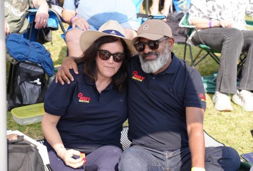 A woman and a man sit close together outdoors on a blanket, both wearing sunglasses and navy blue HOT NORTH shirts. The man has his arm around the woman as they smile, enjoying what looks like a Dave Matthews concert. Other people are in the background.