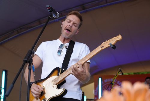 A man in a white t-shirt plays an electric guitar and sings with his eyes closed on an outdoor stage, channeling Dave Matthews as vibrant lights illuminate him and a microphone stands in front.