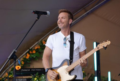 A man in a white t-shirt plays an electric guitar and smiles on an outdoor stage, channeling Dave Matthews vibes. A microphone stands before him and a keyboard with sunflowers brightens the background. Sunglasses hang from his collar.