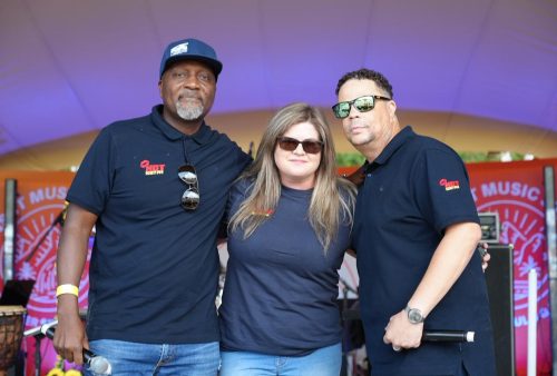 Three people stand together on stage at an outdoor event, wearing matching dark shirts. The two men and the woman in the center—all sporting sunglasses—pose confidently, with colorful event banners and a Dave Matthews sign visible in the background.