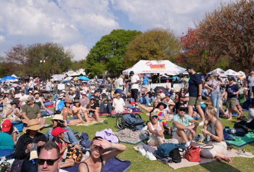 A large crowd of people sit and relax on blankets at an outdoor festival in a park, with trees, tents, and a partly cloudy sky in the background. Many attendees wear hats and sunglasses, enjoying live music reminiscent of a Dave Matthews concert.
