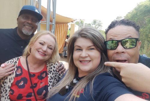 Four adults pose together outdoors, smiling at the camera. One man points at the woman in front. The background shows trees and a yellow building. Everyone appears happy and relaxed, perhaps enjoying a day out after a Dave Matthews concert.