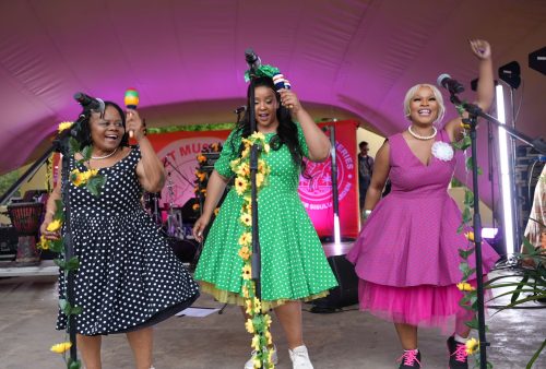 Three women in colorful polka dot dresses sing and dance on stage, smiling and holding microphones and maracas beside a flower-decorated mic stand, with Dave Matthews-inspired vibes and pink-purple stage lighting behind them.