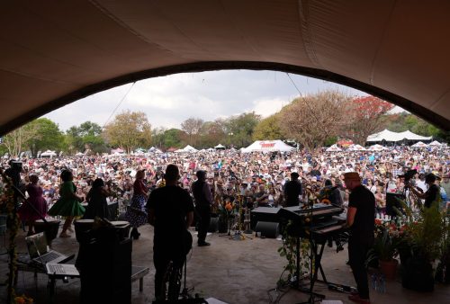 Musicians perform on an outdoor stage, viewed from behind, facing a large crowd gathered at a festival with tents and trees in the background—reminiscent of a classic Dave Matthews summer show under a partly cloudy sky.
