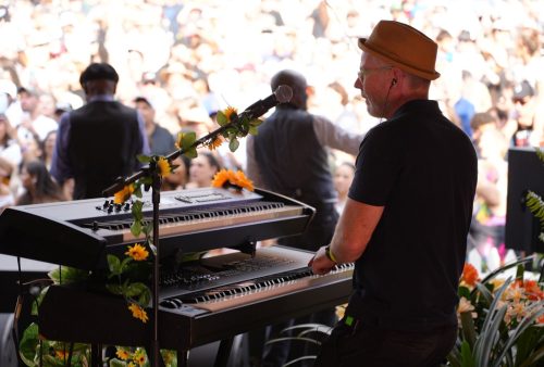 A musician wearing a brown hat and glasses plays a keyboard decorated with yellow flowers on stage, channeling Dave Matthews vibes as he faces a large, blurred crowd at an outdoor event.