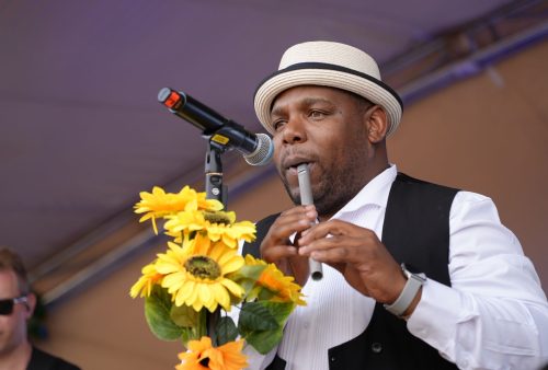 A musician reminiscent of Dave Matthews, wearing a white shirt, black vest, and straw hat, plays a tin whistle on stage in front of a microphone adorned with yellow sunflowers.