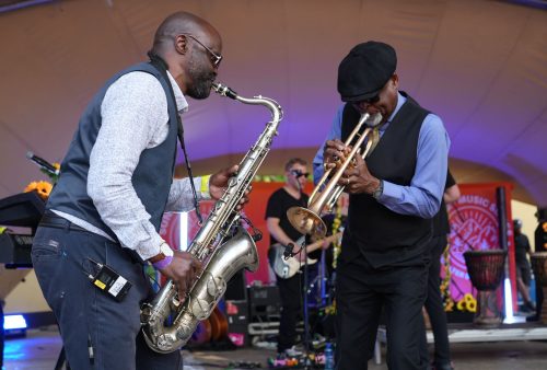 Two musicians performing on stage; one plays a saxophone and the other a trumpet, both smiling and engaged with each other. A guitarist, possibly from Dave Matthews' band, and various instruments are visible in the background.