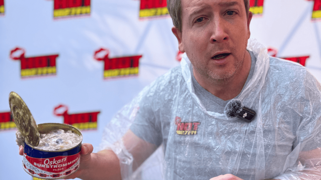 A man wearing a clear plastic poncho holds an open can of Oskars surströmming, making a surprised face during a Teddython segment; a microphone is clipped to his chest and a branded backdrop is behind him.