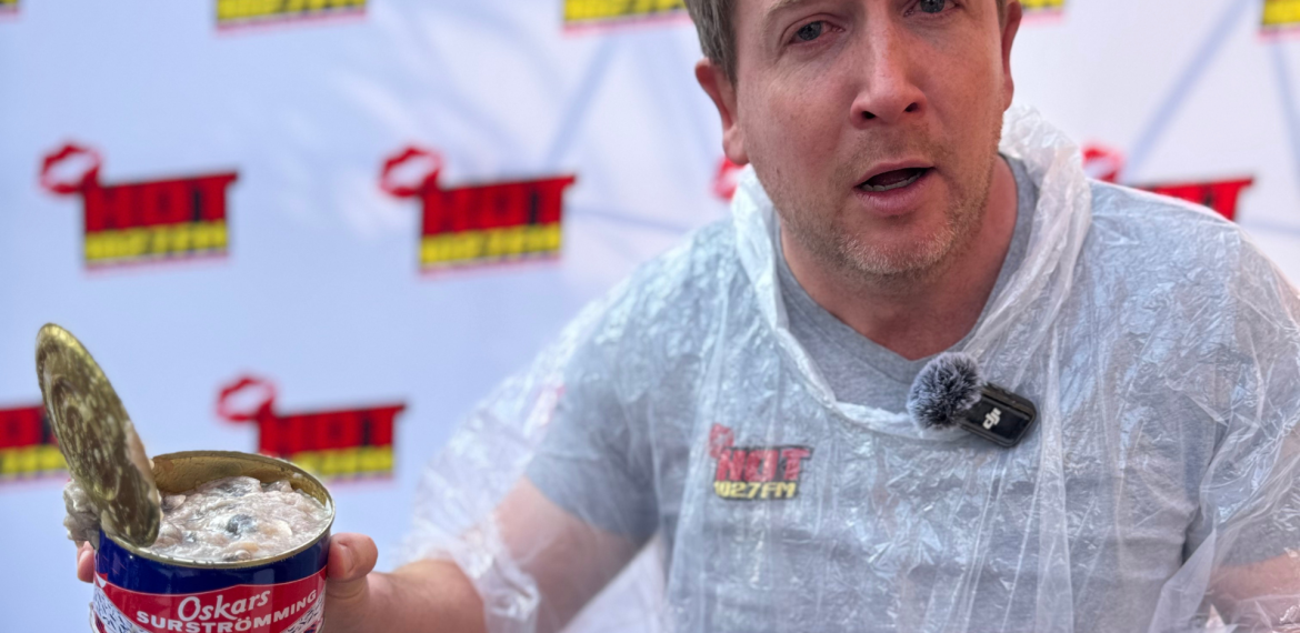 A man wearing a clear plastic poncho holds an open can of Oskars surströmming, making a surprised face during a Teddython segment; a microphone is clipped to his chest and a branded backdrop is behind him.