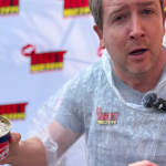 A man wearing a clear plastic poncho holds an open can of Oskars surströmming, making a surprised face during a Teddython segment; a microphone is clipped to his chest and a branded backdrop is behind him.