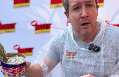 A man wearing a clear plastic poncho holds an open can of Oskars surströmming, making a surprised face during a Teddython segment; a microphone is clipped to his chest and a branded backdrop is behind him.