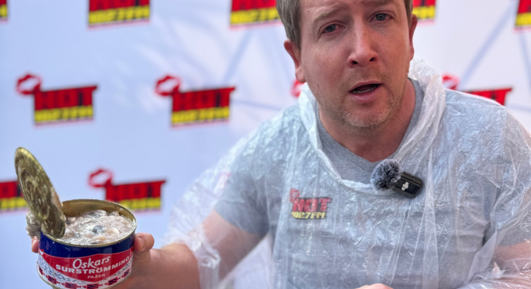 A man wearing a clear plastic poncho holds an open can of Oskars surströmming, making a surprised face during a Teddython segment; a microphone is clipped to his chest and a branded backdrop is behind him.