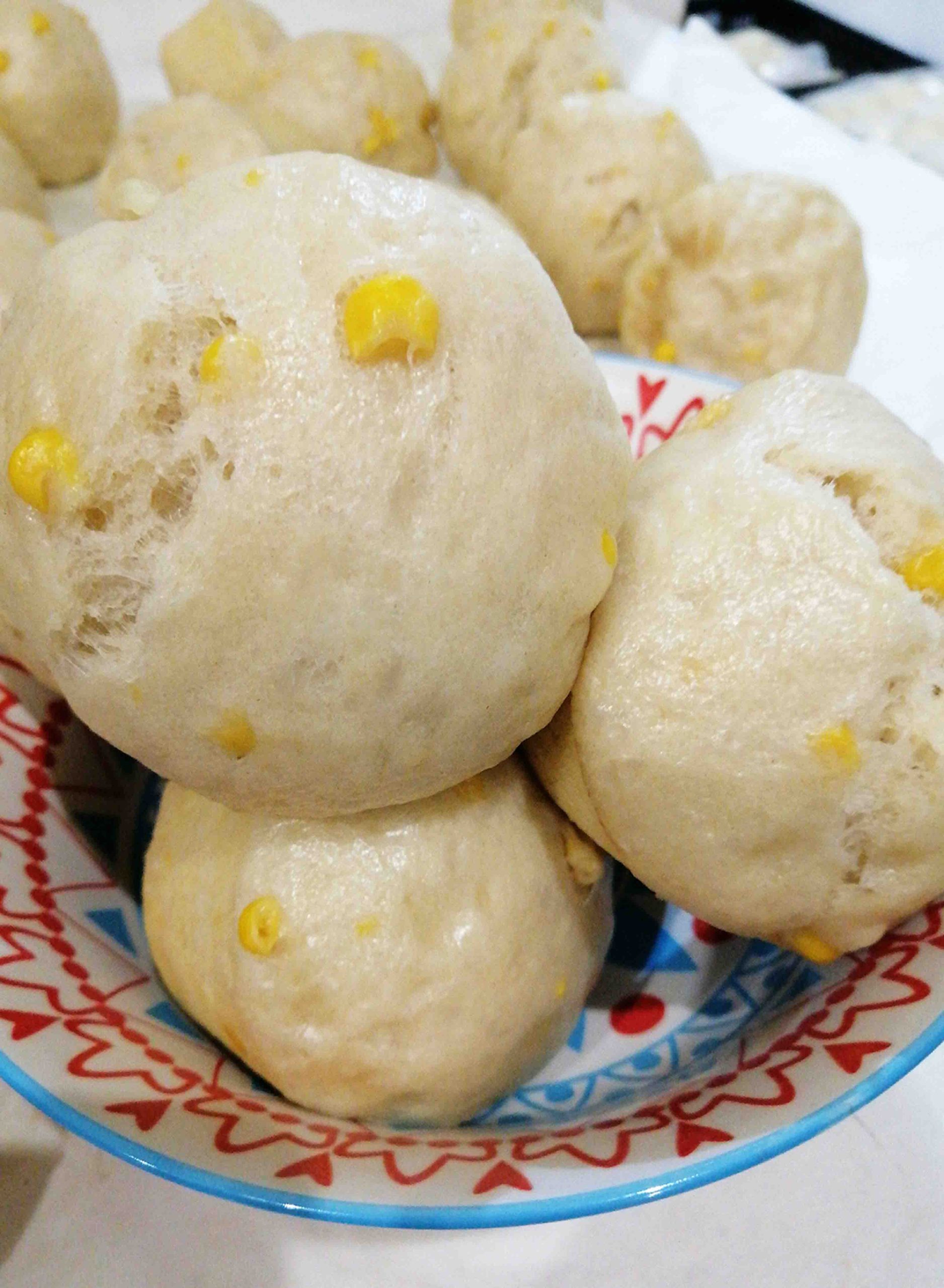 A colorful patterned bowl filled with soft, steamed buns, featuring sweet corn kernels mixed into the dough—a foodie’s delight. More buns are visible in the background on a white surface.