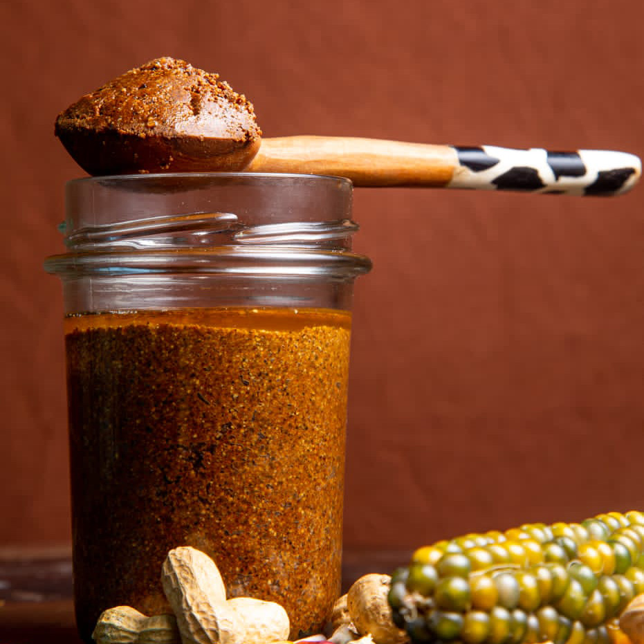 A glass jar filled with a coarse, brown spice mixture sits beside whole peanuts and a multicolored corn cob. Perfect for any foodie, a wooden spoon with a patterned handle rests on the jar, displaying a portion of this unique spice blend.