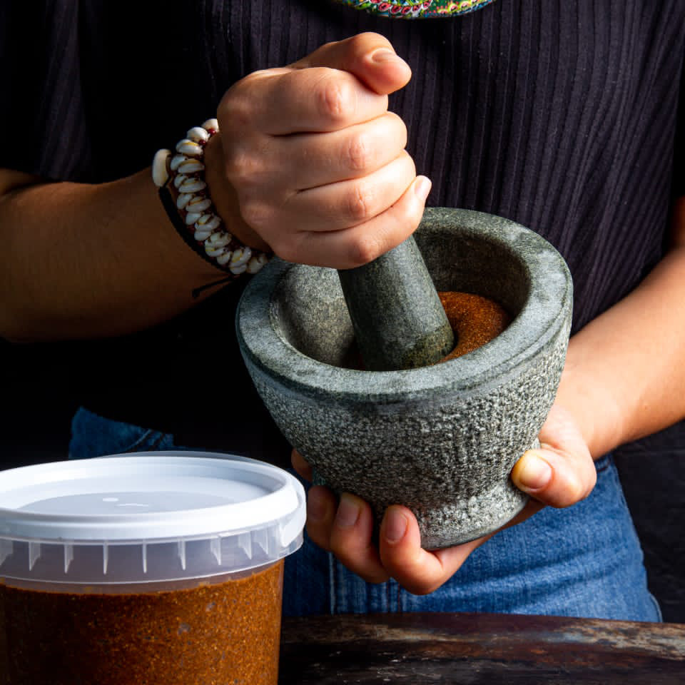 A foodie in a dark shirt uses a stone mortar and pestle to grind spices. Their left hand holds the mortar while the right presses the pestle, with a container of ground spice nearby on the table.