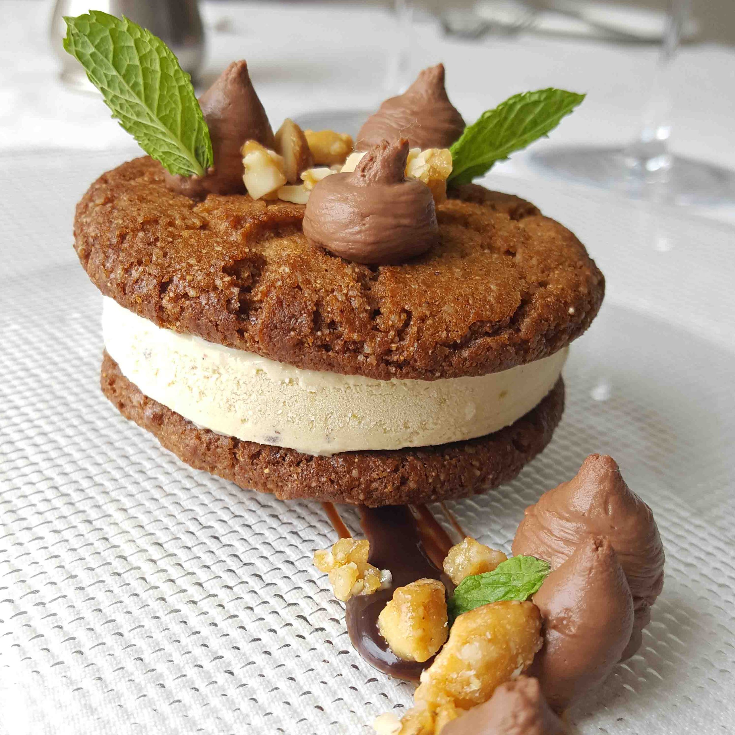 A foodie favorite: two chocolate cookies sandwiching creamy ice cream, topped with chocolate cream dollops, nuts, and mint leaves, served on a white plate drizzled with chocolate sauce.