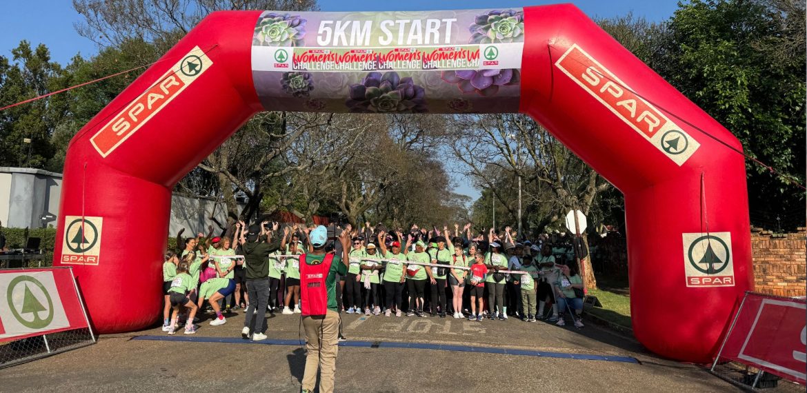 A large group of people in green shirts gathers under a red inflatable arch marked 5KM START at the SPAR Women’s Challenge, with a photographer capturing the moment. Trees and blue sky are visible in the background.