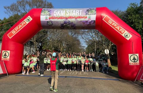 A large group of people in green shirts gathers under a red inflatable arch marked 5KM START at the SPAR Women’s Challenge, with a photographer capturing the moment. Trees and blue sky are visible in the background.