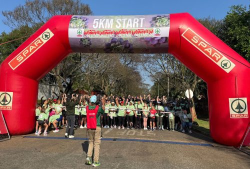 A large group of people in green shirts gathers under a red inflatable arch marked 5KM START at the SPAR Women’s Challenge, with a photographer capturing the moment. Trees and blue sky are visible in the background.