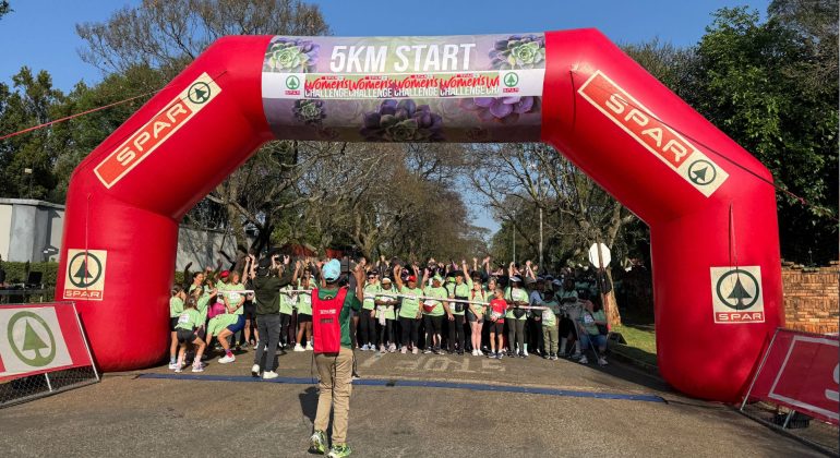 A large group of people in green shirts gathers under a red inflatable arch marked 5KM START at the SPAR Women’s Challenge, with a photographer capturing the moment. Trees and blue sky are visible in the background.