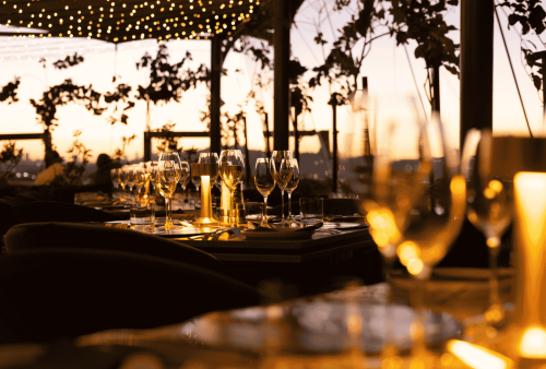 A warmly lit outdoor restaurant at sunset, with tables set for Heritage Day celebrations. Wine glasses, plates, and candles gleam under string lights, creating a cozy and elegant atmosphere surrounded by lush plants.