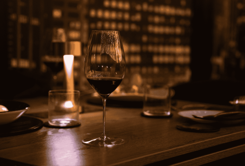 A dimly lit restaurant table set for two, featuring a glass of red wine, a candle, plates, bowls, and utensils—perfect for a cozy Heritage Day celebration—with a blurred background of wine bottles on shelves.