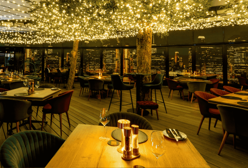 A modern restaurant with floor-to-ceiling windows overlooks the city skyline at night. Warm string lights hang above elegant wooden tables and stylish chairs, creating a cozy, upscale atmosphere—perfect for a memorable Heritage Day celebration.