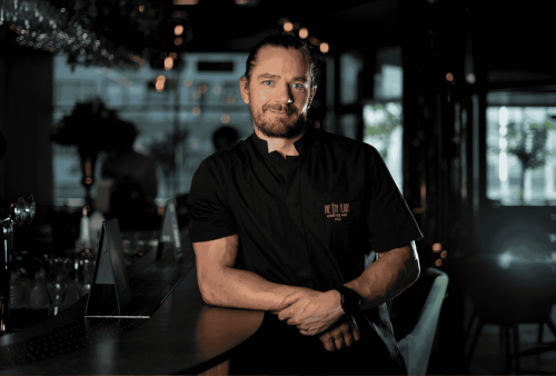 A man with a beard and tied-back hair, wearing a black shirt, leans casually on a bar counter in a dimly lit, modern restaurant—its stylish decor adding to the ambiance, perfect for a relaxed Heritage Day celebration.