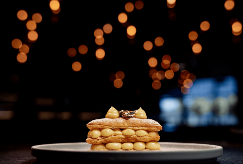 A plated pastry dessert with layers of puff pastry and cream sits centered on a plate, with warm bokeh lights in the dark background, creating an elegant, moody atmosphere—perfect for celebrating Heritage Day in style.