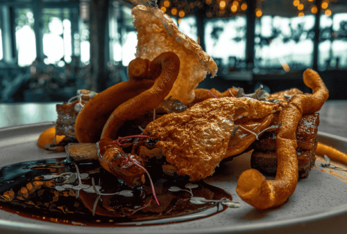 A close-up of an artfully plated gourmet dish, inspired by Heritage Day, featuring crispy fried elements, a glossy dark sauce, and microgreens on a gray plate, with a softly lit restaurant interior in the background.