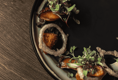 A close-up of a gourmet dish on a dark plate, featuring seared fish, crispy onion rings, microgreens, and artful garnishes—perfect for celebrating Heritage Day on a rustic wooden table.