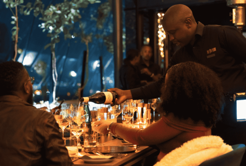 A server pours wine for two people seated at a candlelit restaurant table set with glasses and plates, creating a warm and elegant atmosphere perfect for celebrating heritage day.