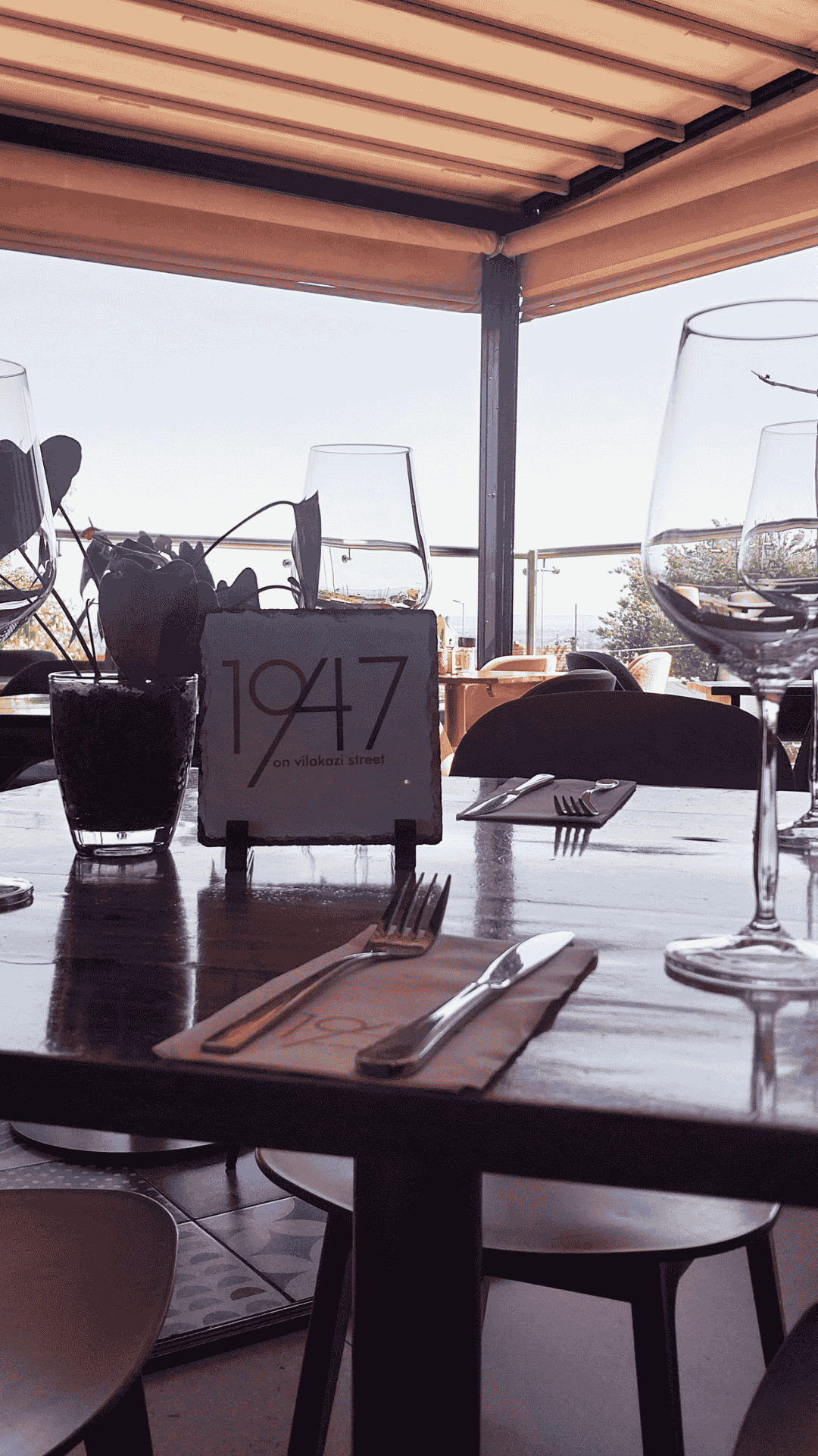 A sunlit restaurant table set with wine glasses, cutlery, a small plant, and a sign reading 1947 on Vilakazi Street invites you to celebrate Heritage Day, with outdoor seating visible in the background.