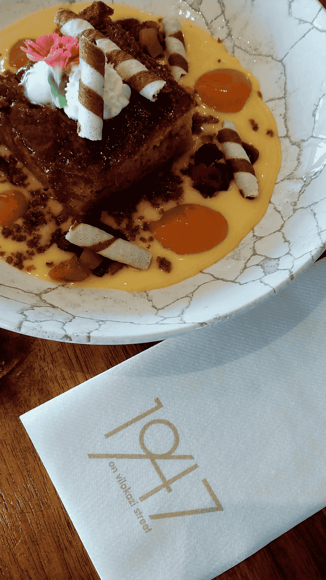 A dessert topped with whipped cream, a pink flower, and rolled wafers sits in a creamy sauce with orange dollops on a marbled plate; perfect for celebrating Heritage Day at 1947 on Waverley Street, as shown on the nearby napkin.