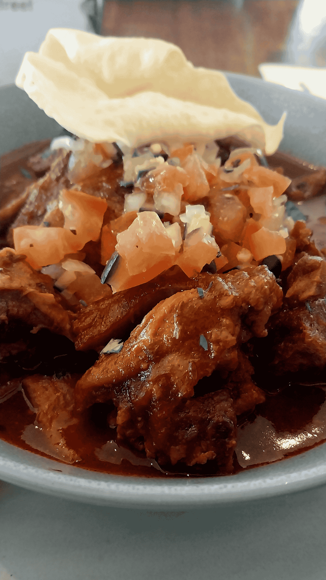 A close-up of a savory meat dish in rich brown sauce, topped with diced tomatoes and onions, served with a crisp, pale cracker on the side. Perfect for celebrating Heritage Day, the dish is beautifully presented on a gray plate.