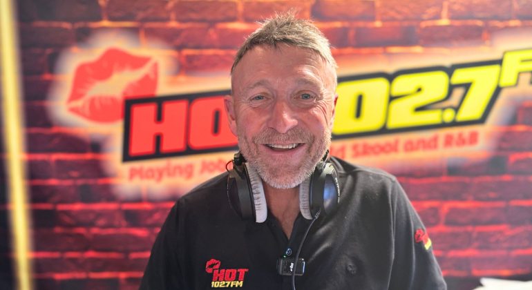 A smiling man wearing headphones and a black shirt with a HOT 102.7 FM logo stands in front of a radio station backdrop featuring the same logo and a brick pattern, ready to discuss topics like music and homeownership on air.