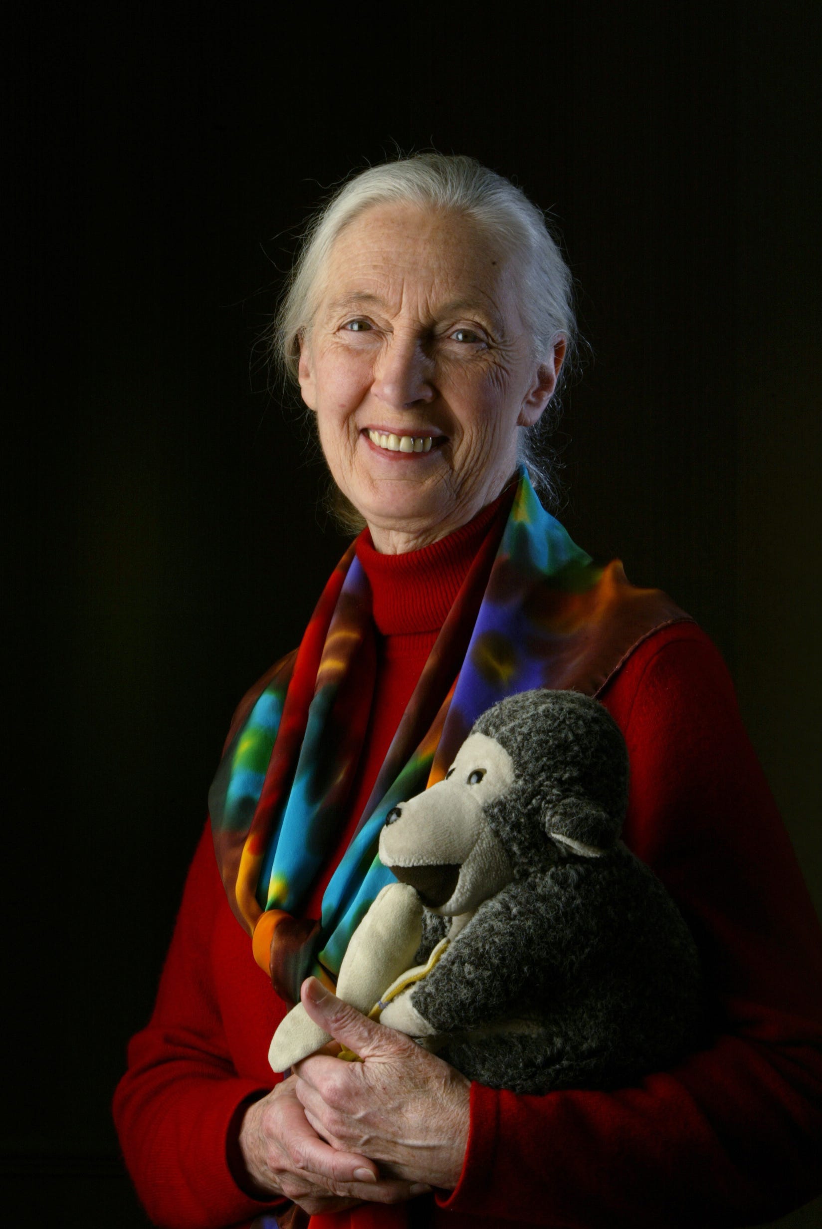 An elderly woman with white hair in a ponytail, wearing a red top and a colorful scarf, smiles joyfully while holding a gray stuffed monkey—her lighthearted expression is the perfect antidote to any brainrot against a dark background.