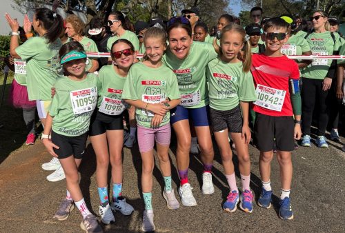 A group of five children and one adult, all wearing green race shirts and numbered bibs, smile and pose together at the SPAR Womens Challenge with other participants in the background.