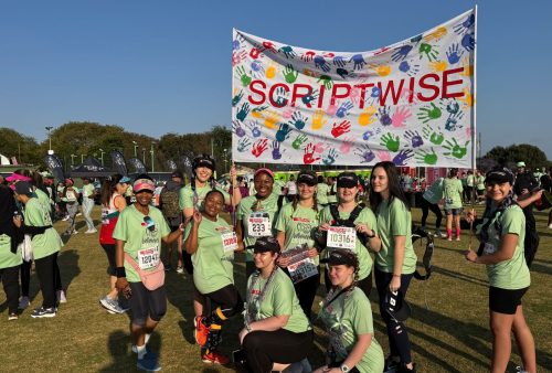A group of people in matching green shirts and race bibs pose in front of a colorful “SCRIPTWISE” banner with handprints at the SPAR Womens Challenge, surrounded by other participants on a grassy field.