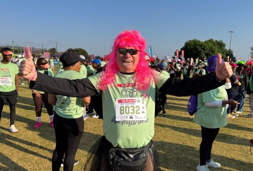 A smiling runner in a bright pink wig, sunglasses, and a tutu gives thumbs up at the crowded SPAR Womens Challenge. Other participants in race bibs and green shirts stand in the background, adding to the festive outdoor atmosphere.