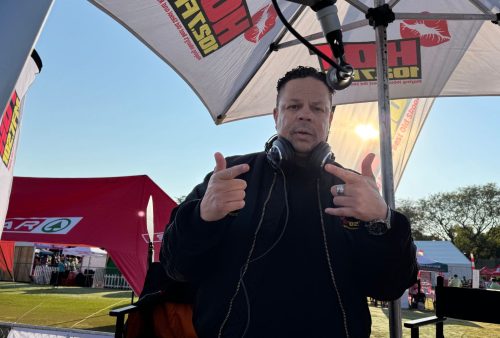 A man with headphones around his neck stands under a promotional umbrella at the SPAR Women's Challenge outdoor event, pointing towards the camera. Red tents and a grassy area with people are visible in the background.