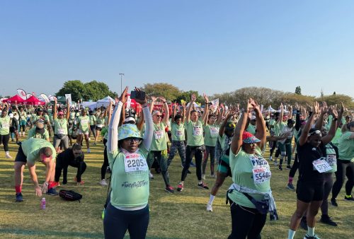 A large group of people in green shirts and race bibs are stretching and warming up on a grassy field before the SPAR Womens Challenge, with tents and trees in the background under a clear blue sky.